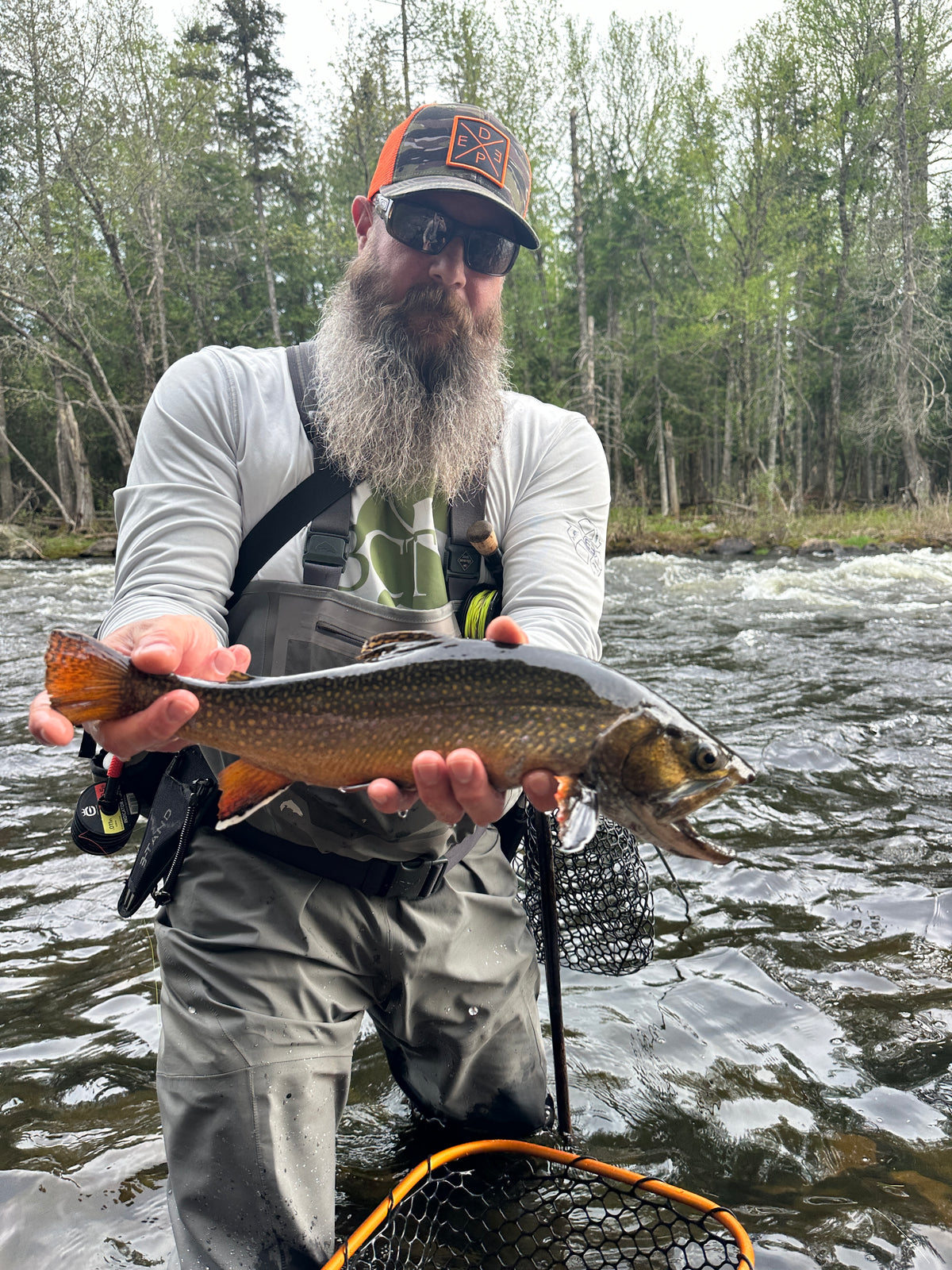 Koby holding a large brookie  by a river in maine with trees in the background